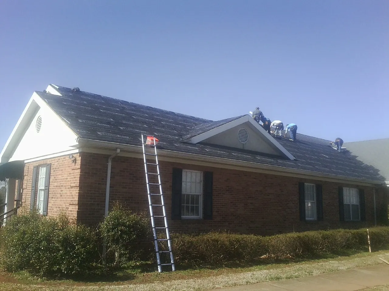 Crew tearing shingles off a brick ranch roof with a ladder set — Emergency Roof Repair work by our crew in Ware