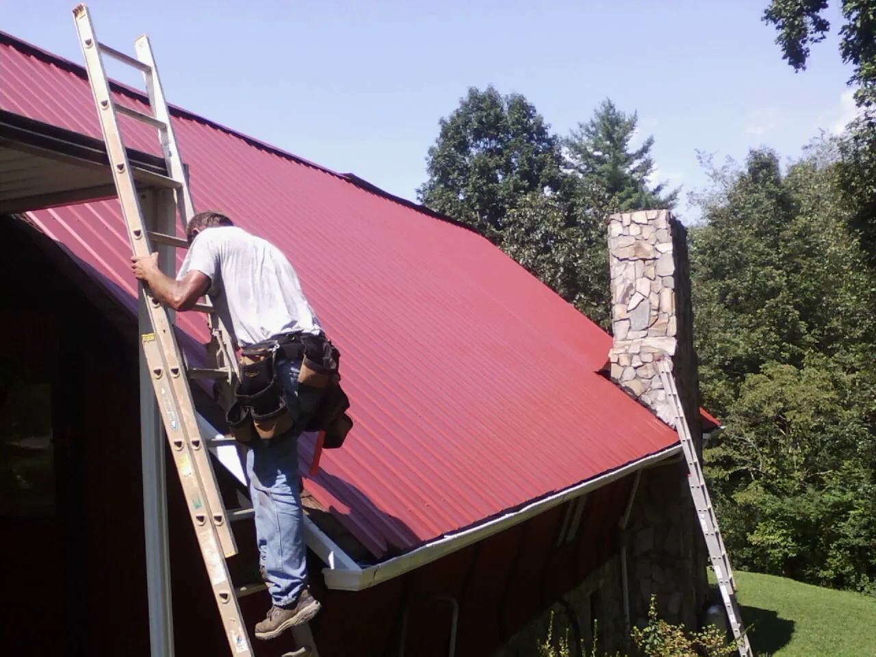 Roofer on a ladder beside a red metal roof and stone chimney — Soffit & Fascia Repair by Roofing Brothers in Ware